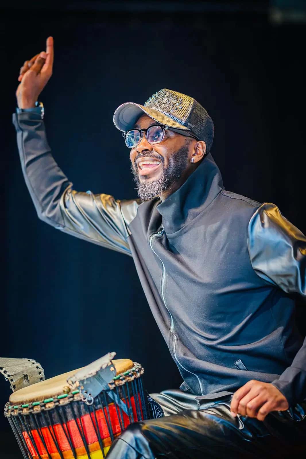 A smiling percussionist raises his hand mid-performance beside a colourful djembe